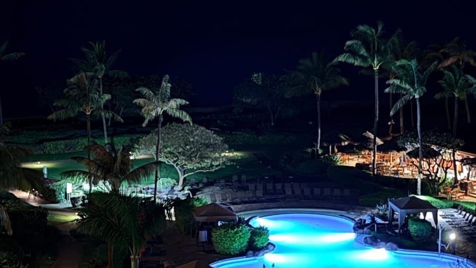 Pre-dawn view of a quiet Maui shoreline with a softly lit pool below and the dark Pacific Ocean beyond.